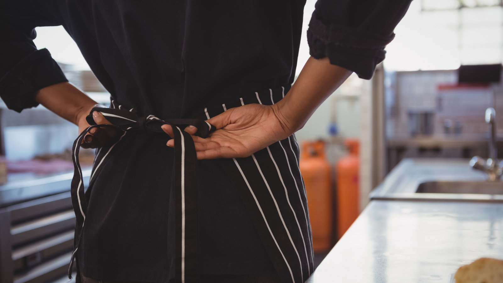 Black woman photographed from behind tying the ties on her striped apron, standing in an industral kitchen.